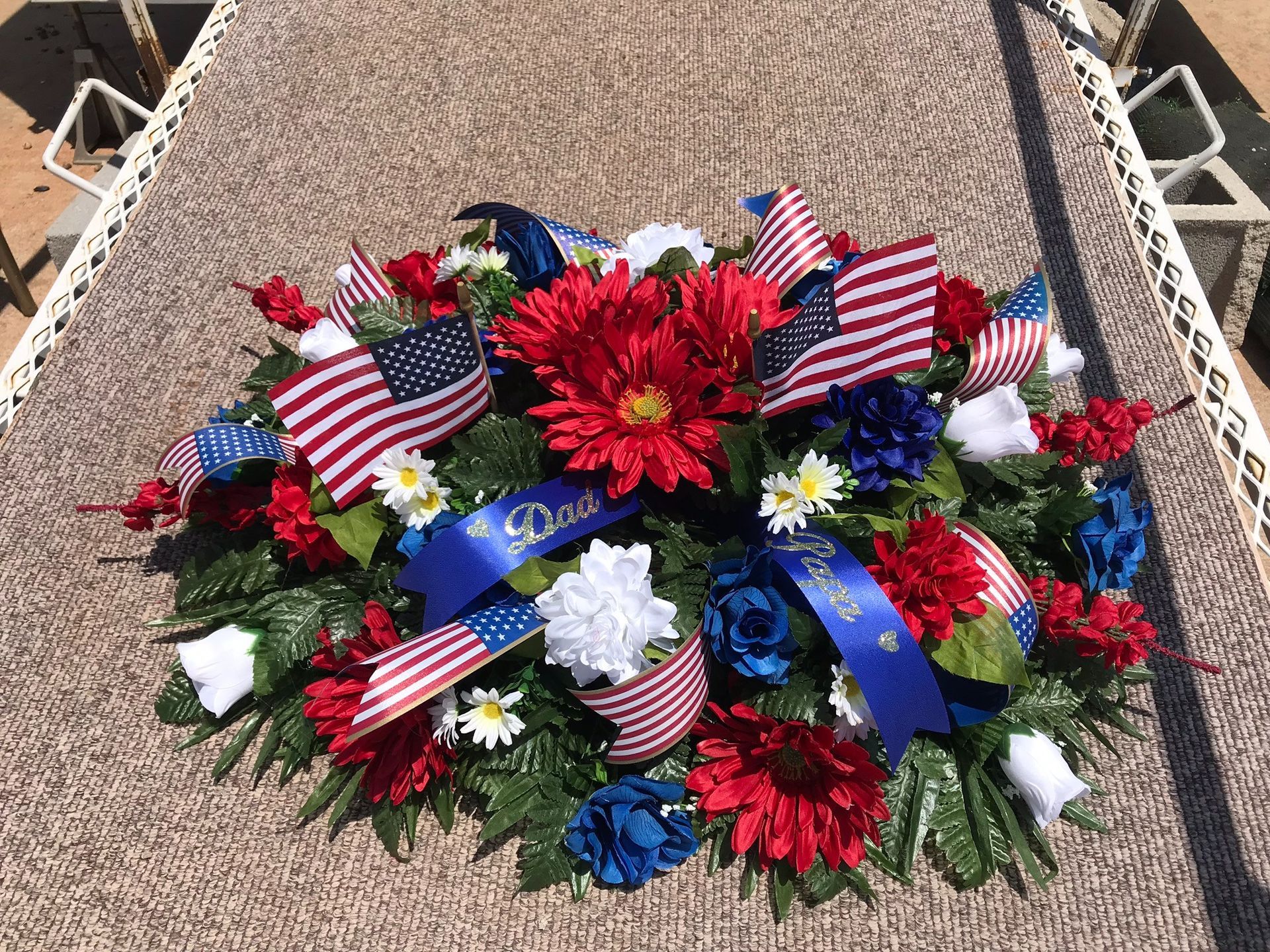 Red, white, and blue memorial wreath with American flags and flowers on a gravel gravesite