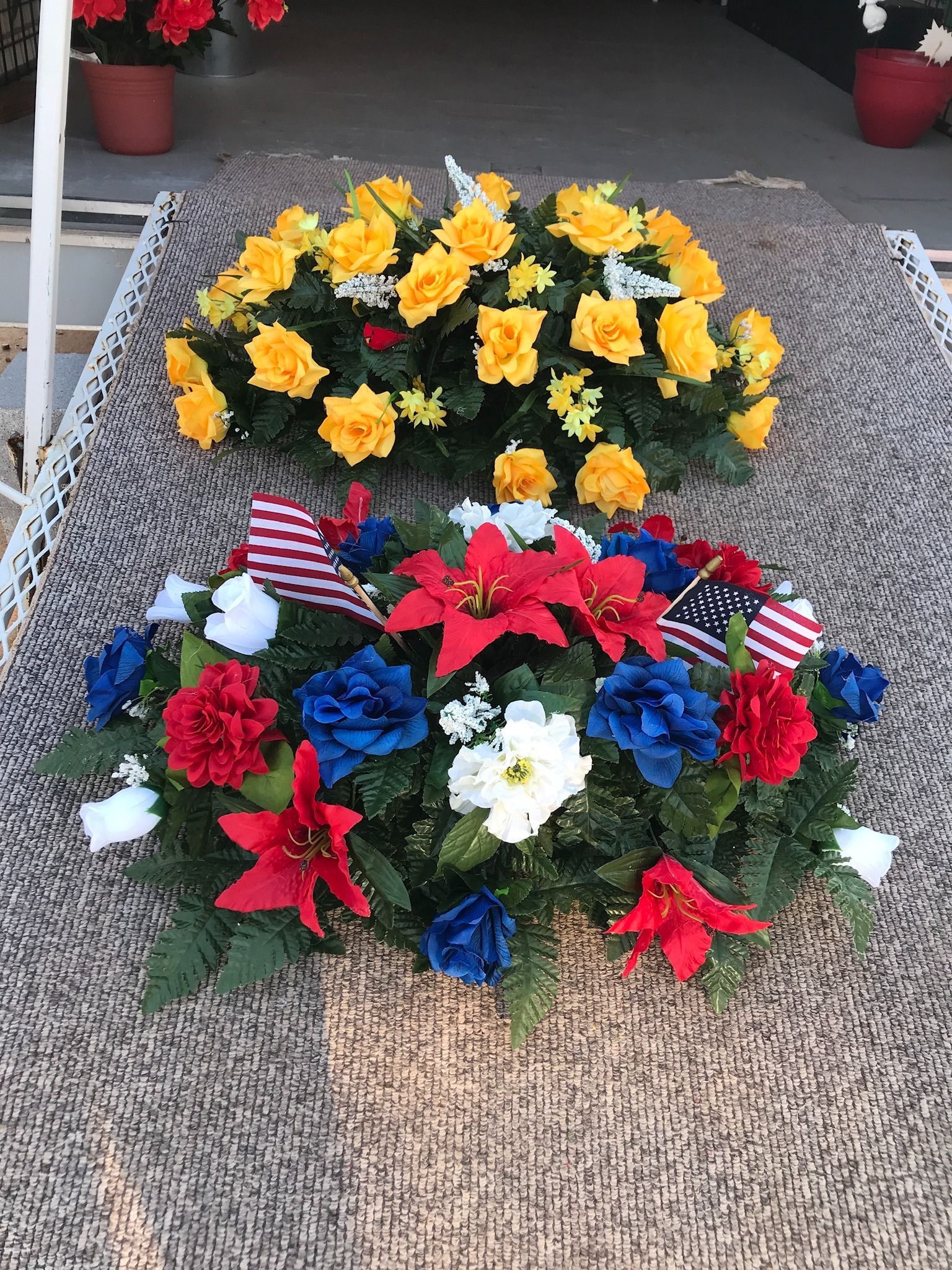 Red, white, and blue floral wreath with small American flags, below a yellow flower arrangement on a porch mat