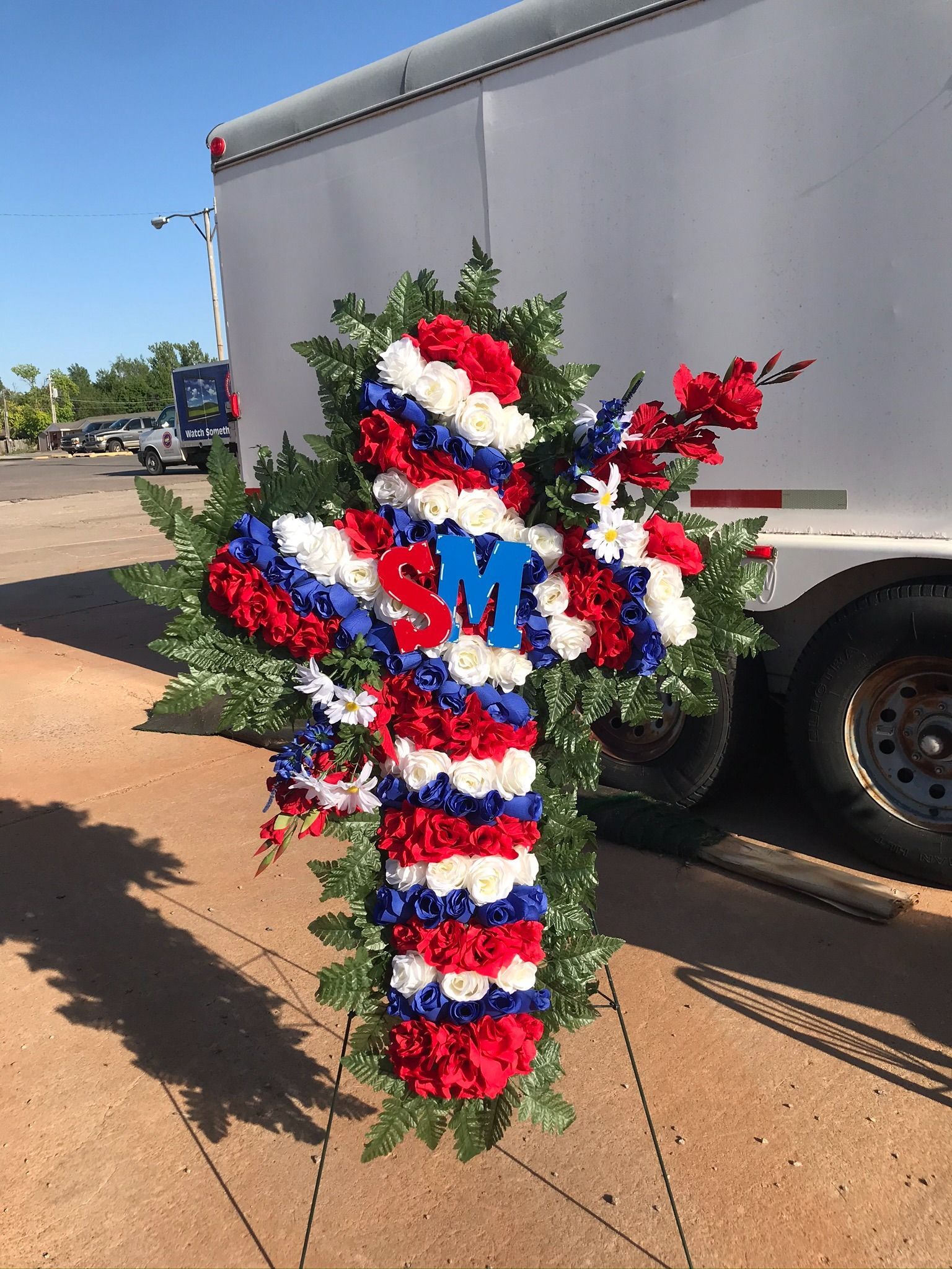 Red, white, and blue floral cross with “SM” on a trailer beside a road
