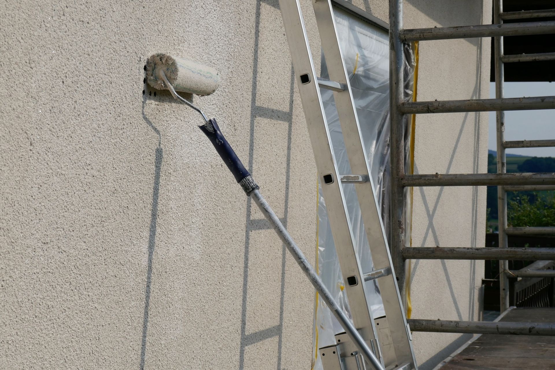 Painter’s roller on an extension pole against a stucco wall beside a ladder and scaffold
