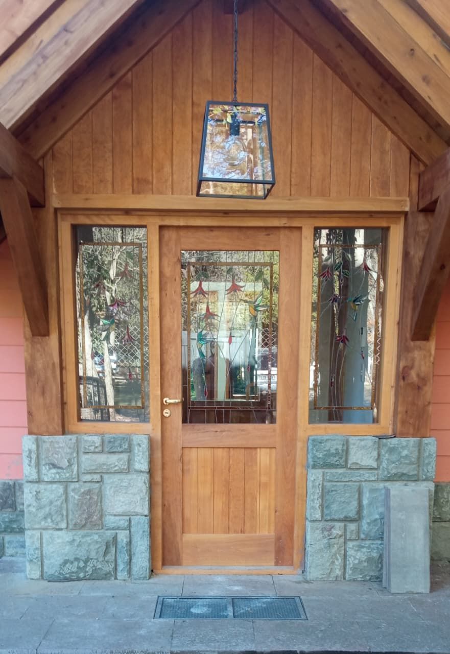 Entrada de cabaña de madera con base de piedra, puerta frontal de cristal y pequeña vidriera en la parte superior.