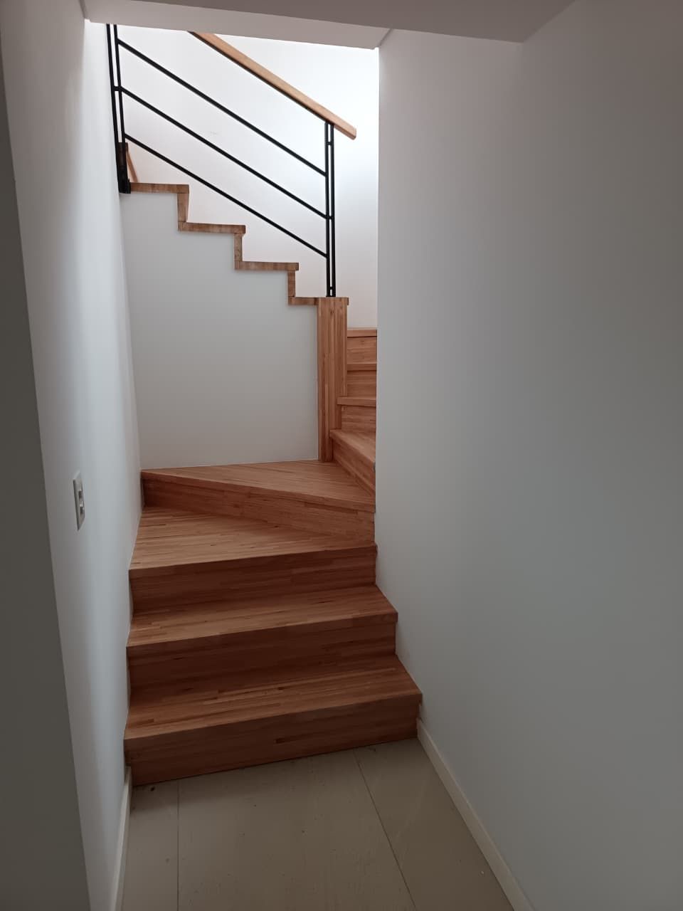 Escalera interior estrecha con escalones de madera, paredes blancas y barandilla de metal negro.