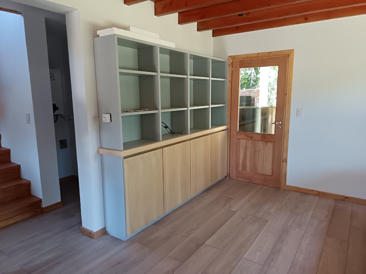 Habitación vacía con suelo de madera, vigas de techo a la vista, vitrina con puerta de cristal y puerta abierta al exterior.
