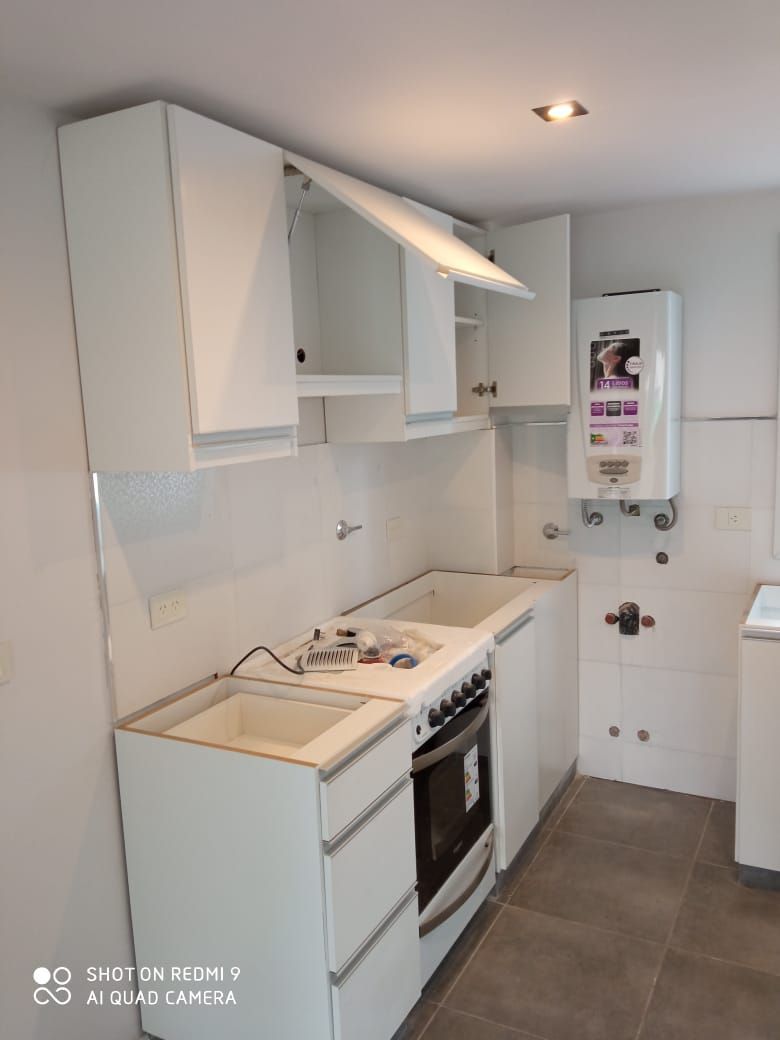Cocina compacta tipo galera de color blanco con estufa, fregadero, gabinetes y piso de baldosas grises.
