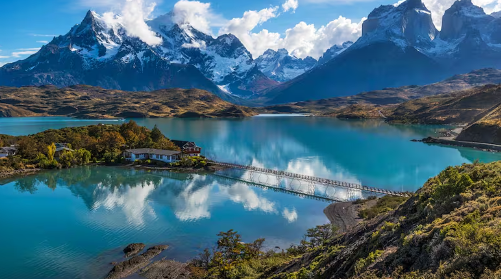 Montañas cubiertas de nieve se alzan imponentes sobre un lago turquesa que refleja el cielo, con un pequeño edificio en una península.