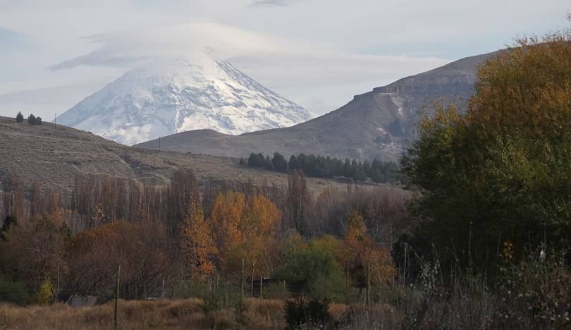 Una montaña cubierta de nieve se alza tras un primer plano de árboles con colores otoñales y colinas onduladas bajo un cielo nublado.