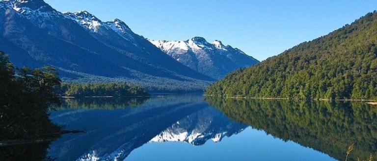 Las montañas nevadas se reflejan en un lago tranquilo y cristalino, flanqueado por densos bosques verdes bajo un cielo azul brillante.