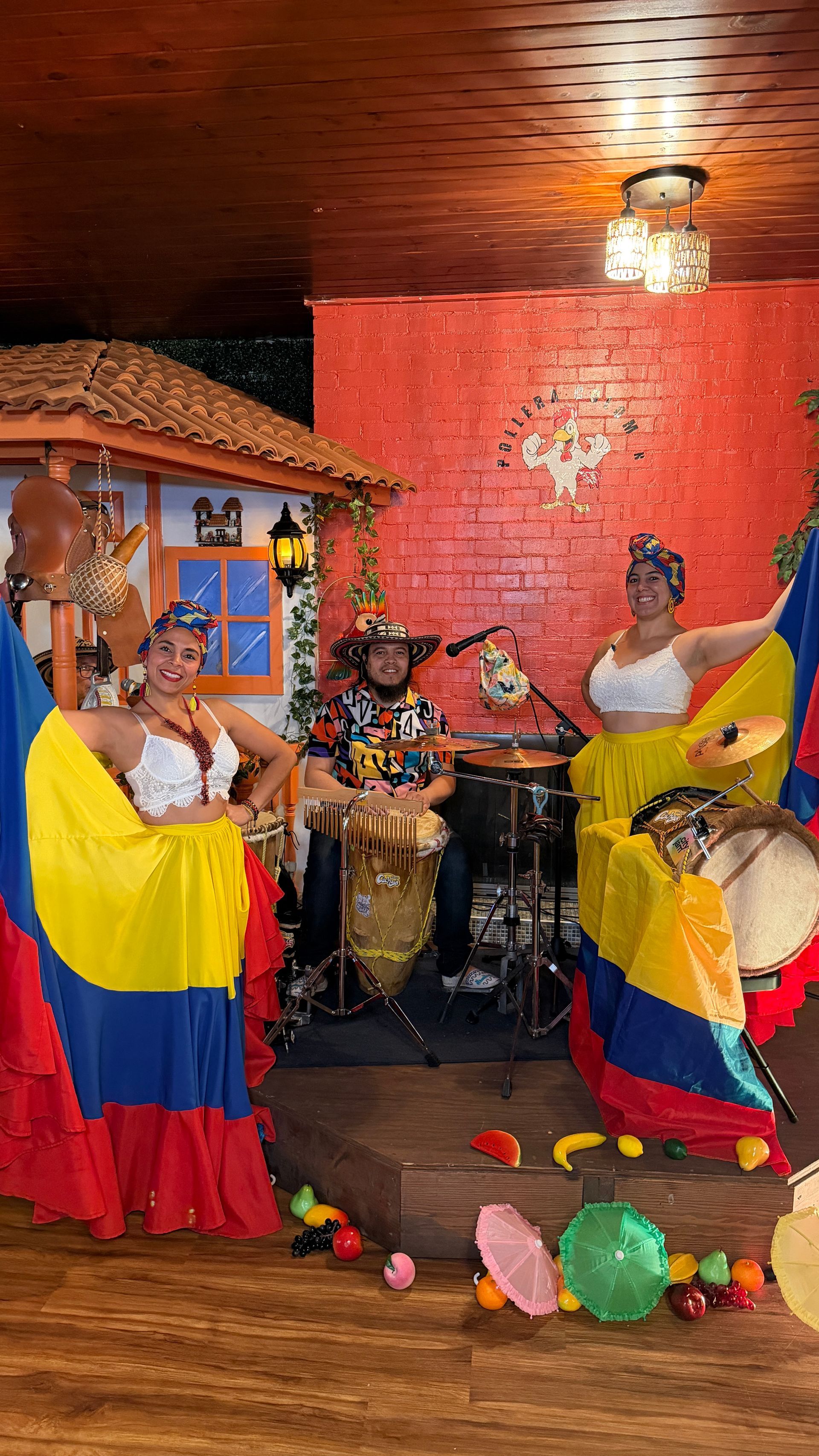 Colombian dancers in colorful dresses perform with a drummer in front of a rustic backdrop.