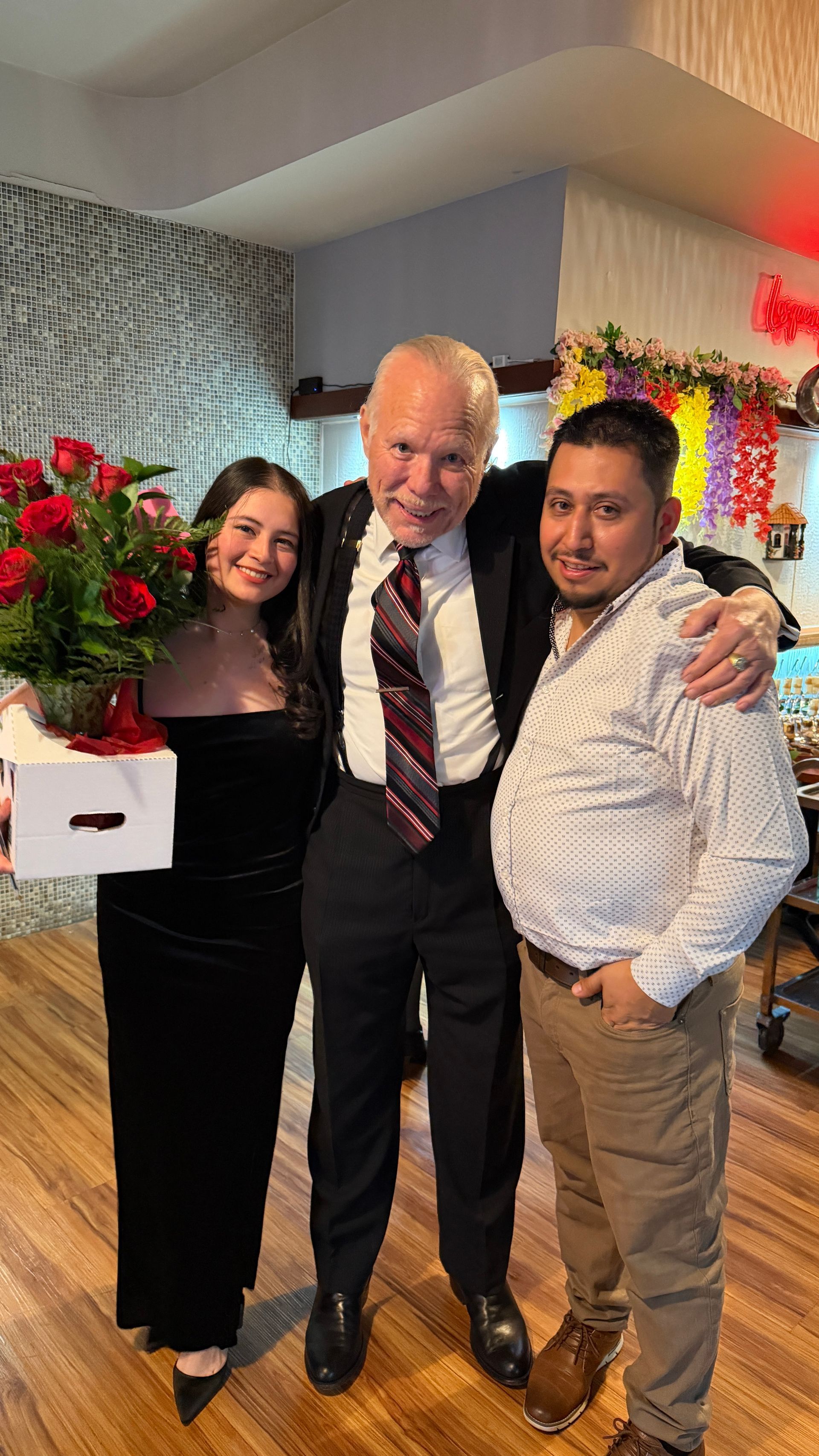 Three people pose for a photo indoors, one holds red roses.