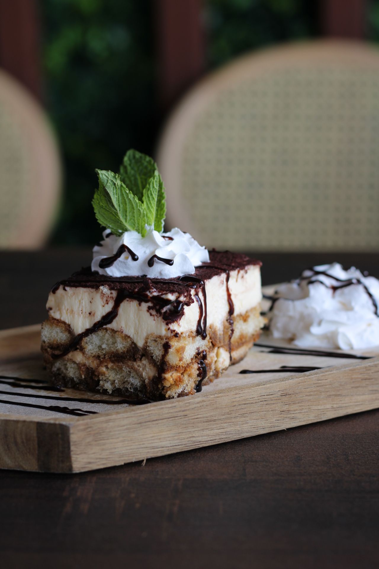Tiramisu dessert on wooden board, drizzled with chocolate and whipped cream, garnished with mint.