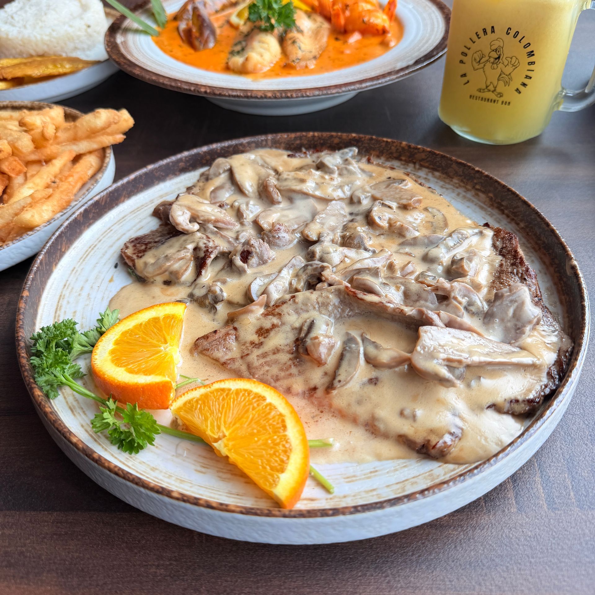 Steak with mushroom sauce on a plate, garnished with orange slices. Other dishes and drink visible.