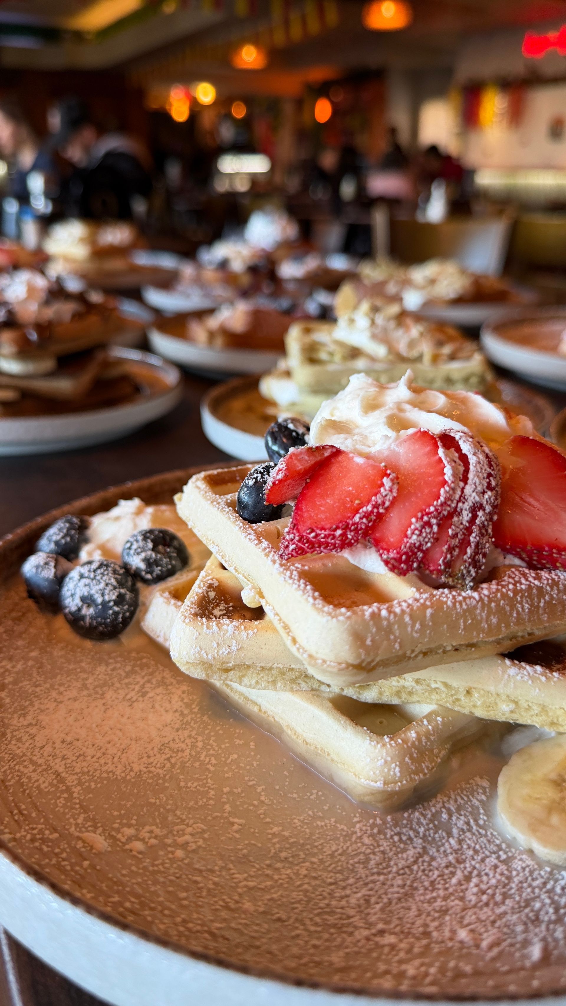 Stack of waffles with strawberries, blueberries, and whipped cream, sprinkled with powdered sugar.