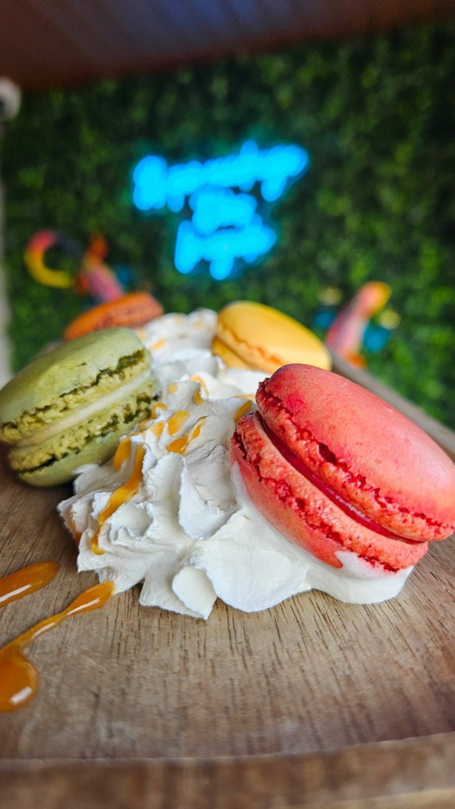 Colorful macarons on whipped cream with caramel drizzle, on a wooden board. Neon sign in background.