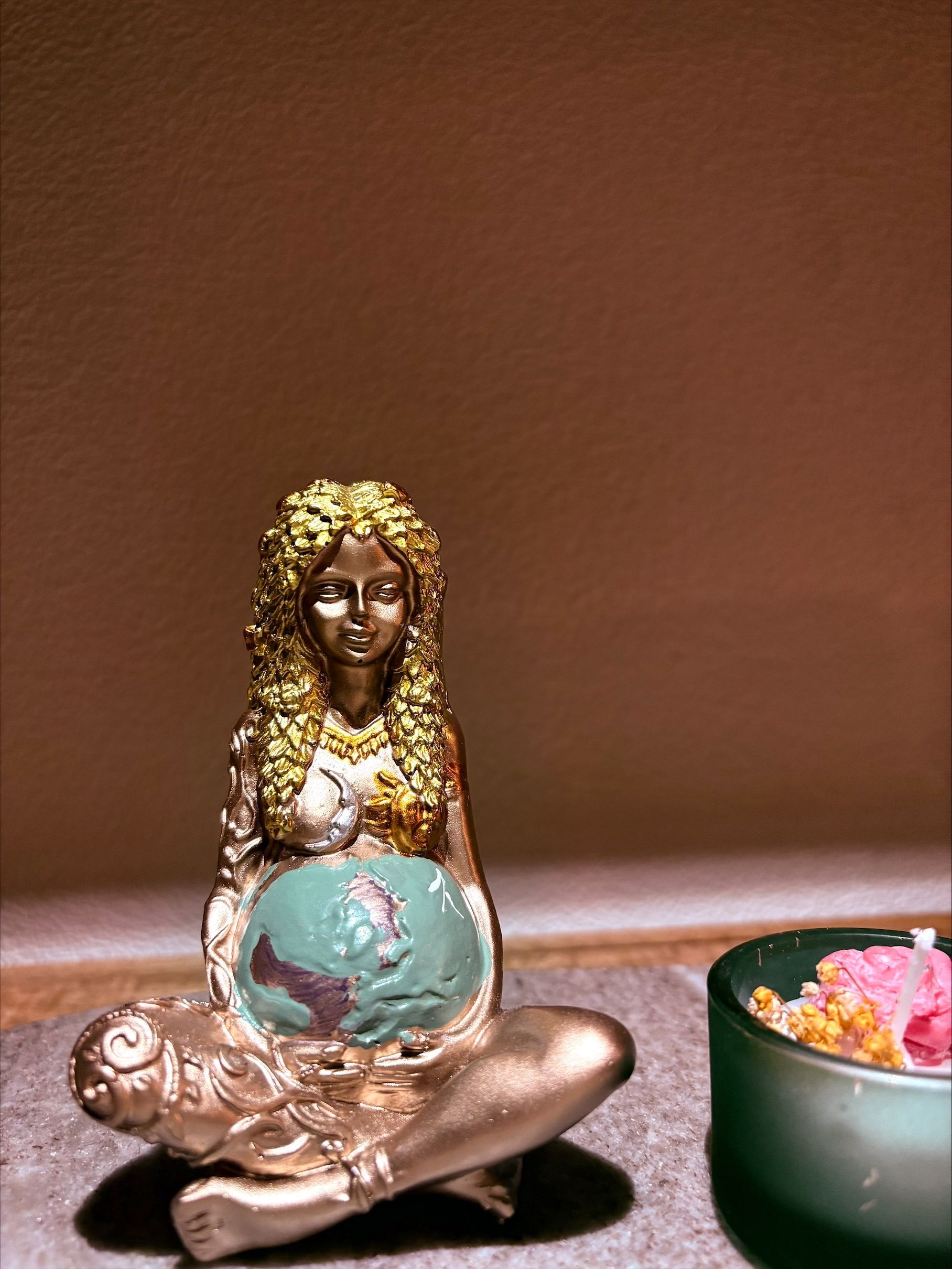 Gouden beeld van een zittende vrouw die een blauwe wereldbol vasthoudt. Ernaast staat een kaars in een groene houder.