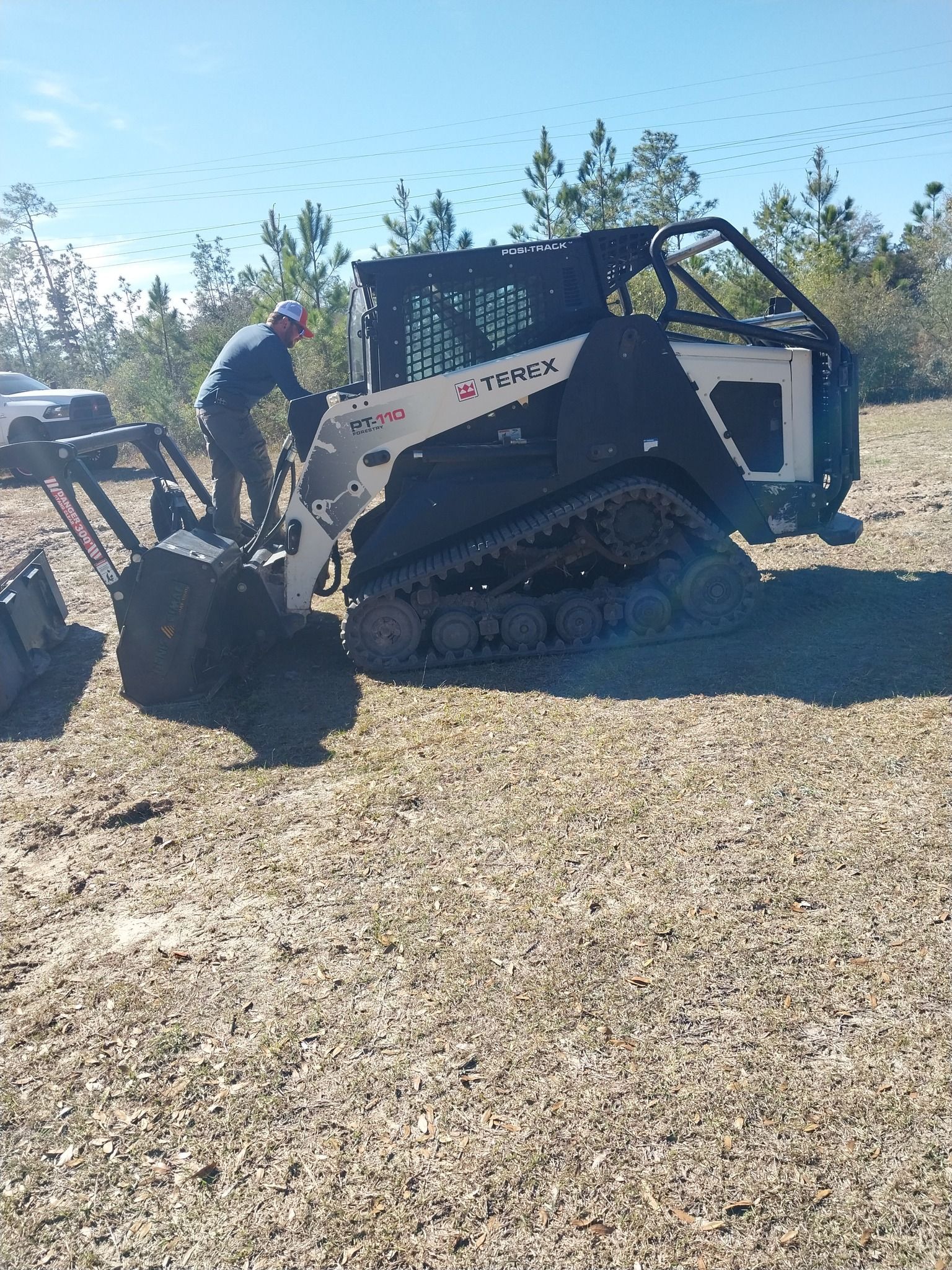 Man operating a tracked skid steer with a forestry cutter attachment in a field on a sunny day.