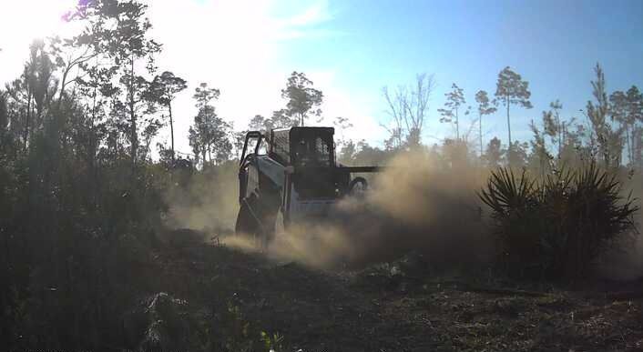 A Bobcat skid-steer stirs up dust while clearing brush in a field under a sunny sky.