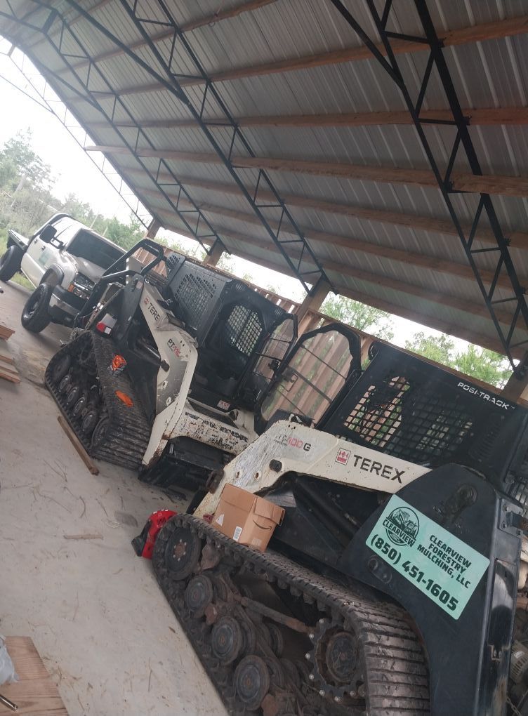 Skid steer track loader parked under a metal roof, next to a pickup truck.