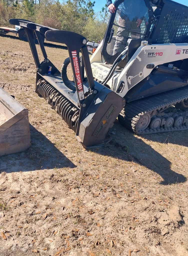 Skid steer with mulching head on dry dirt. Operator inside. Outdoors, sunny day.