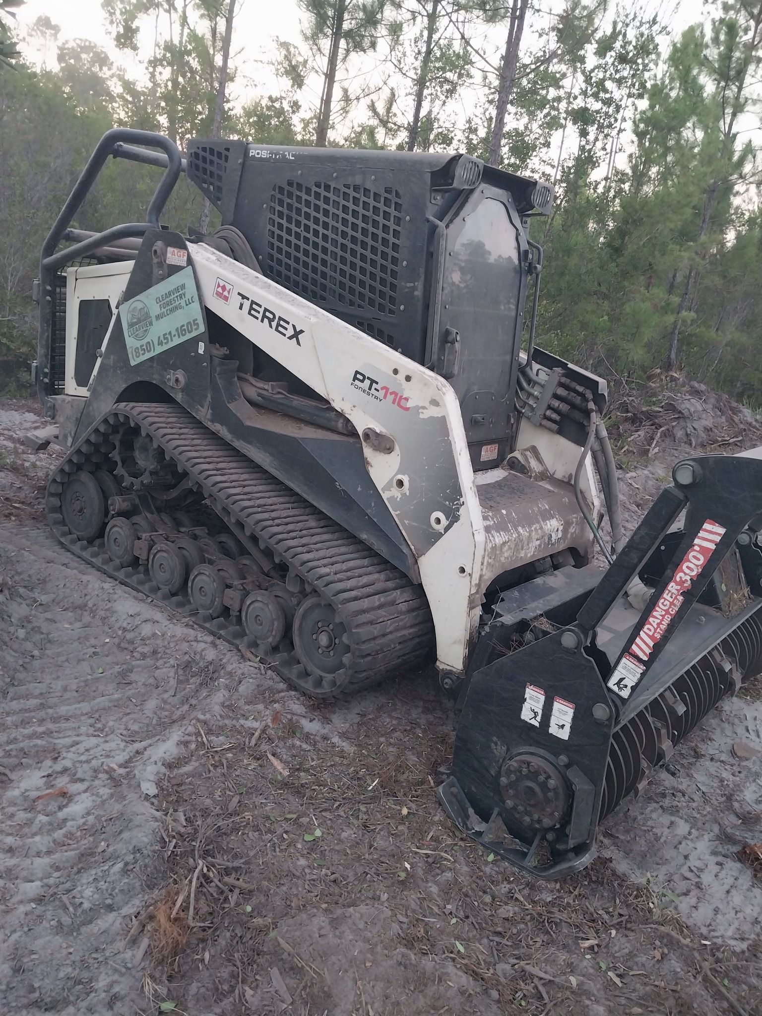 Skid steer with mulching head on dirt path, surrounded by trees.