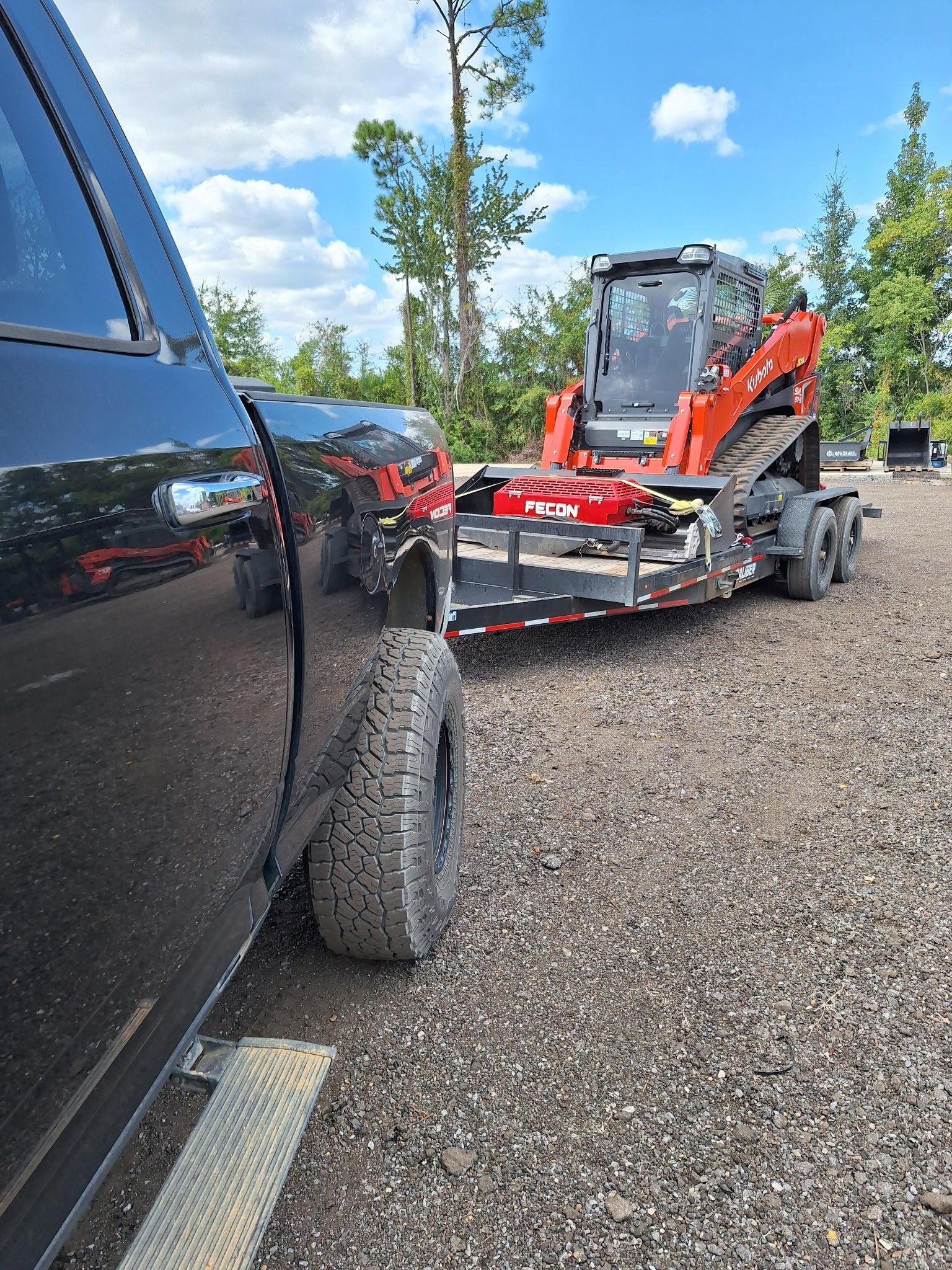 Black truck towing a trailer with an orange and black track skid steer on gravel. Sunny day.