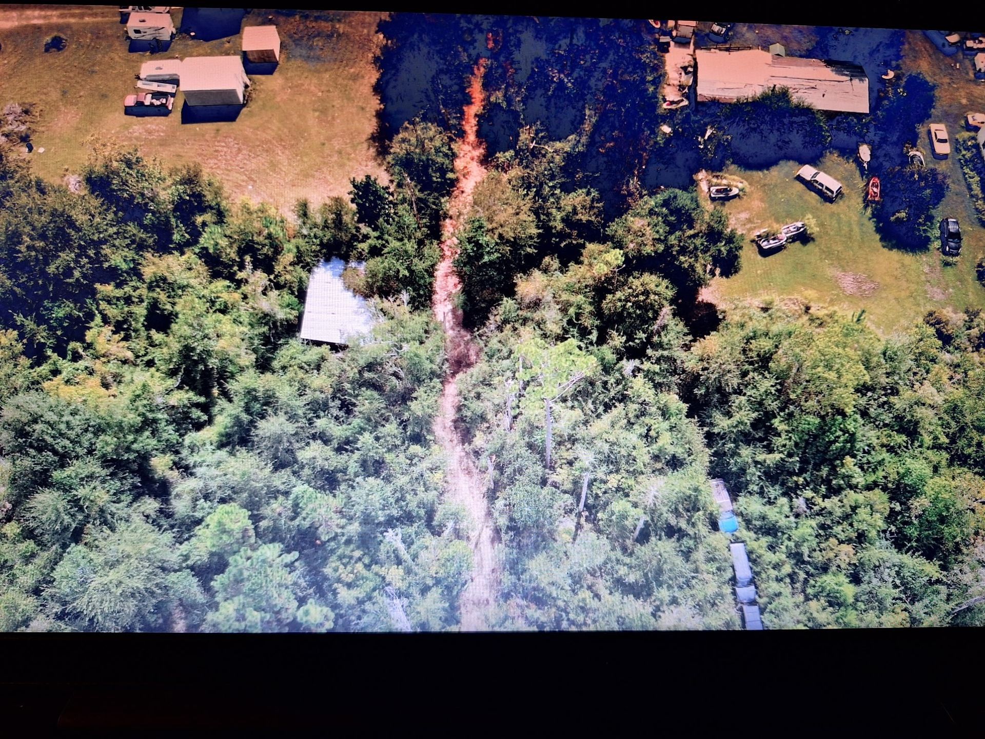 Overhead view of a dirt path through dense green trees, flanked by structures and vehicles on either side in a rural area.