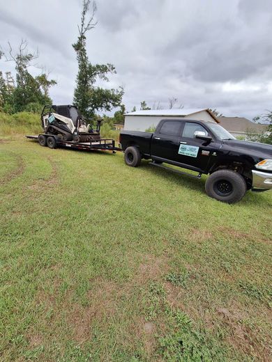Black pickup truck towing a trailer with landscaping equipment on a grassy field under a cloudy sky.