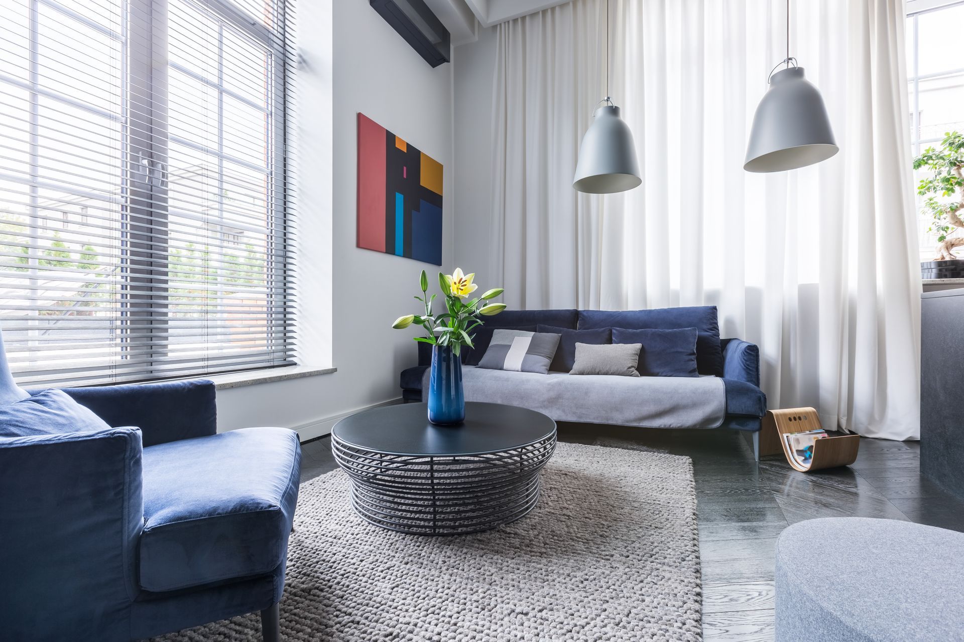 Modern living room with blue sofa, armchair, round coffee table, and large windows with sheer curtains