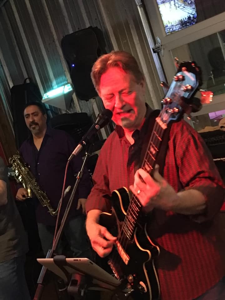 A man is playing a guitar and singing into a microphone while another man plays a saxophone.