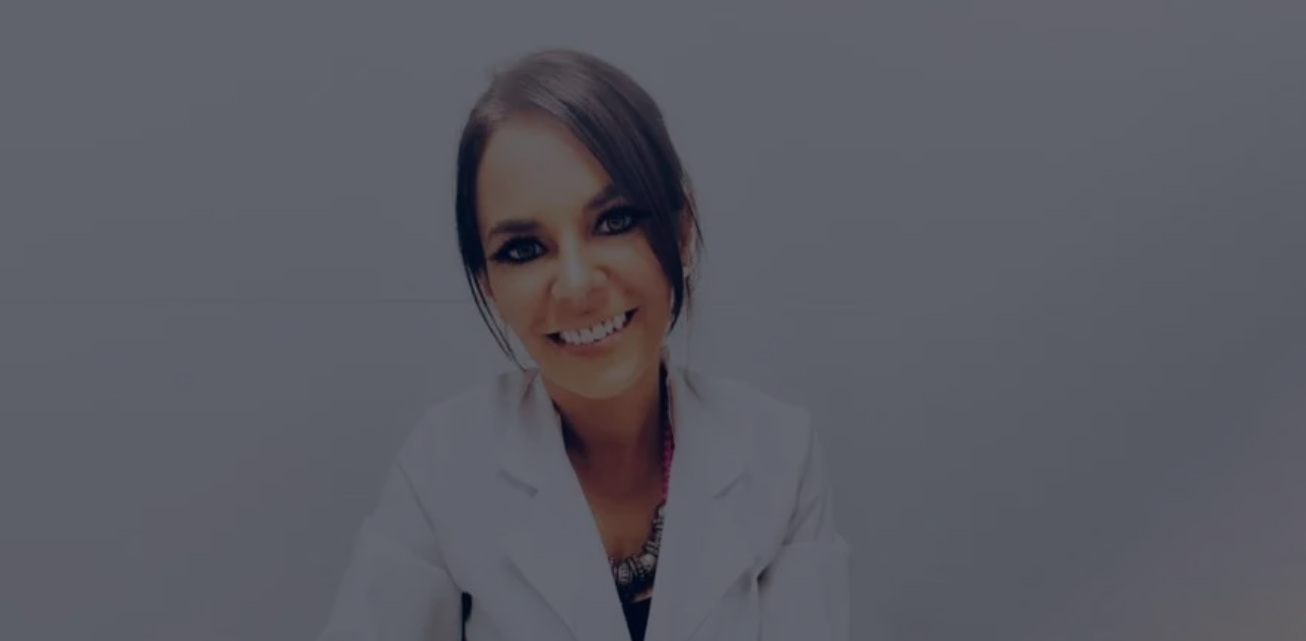 Mujer con bata blanca de laboratorio sonríe a la cámara. Fondo gris neutro.