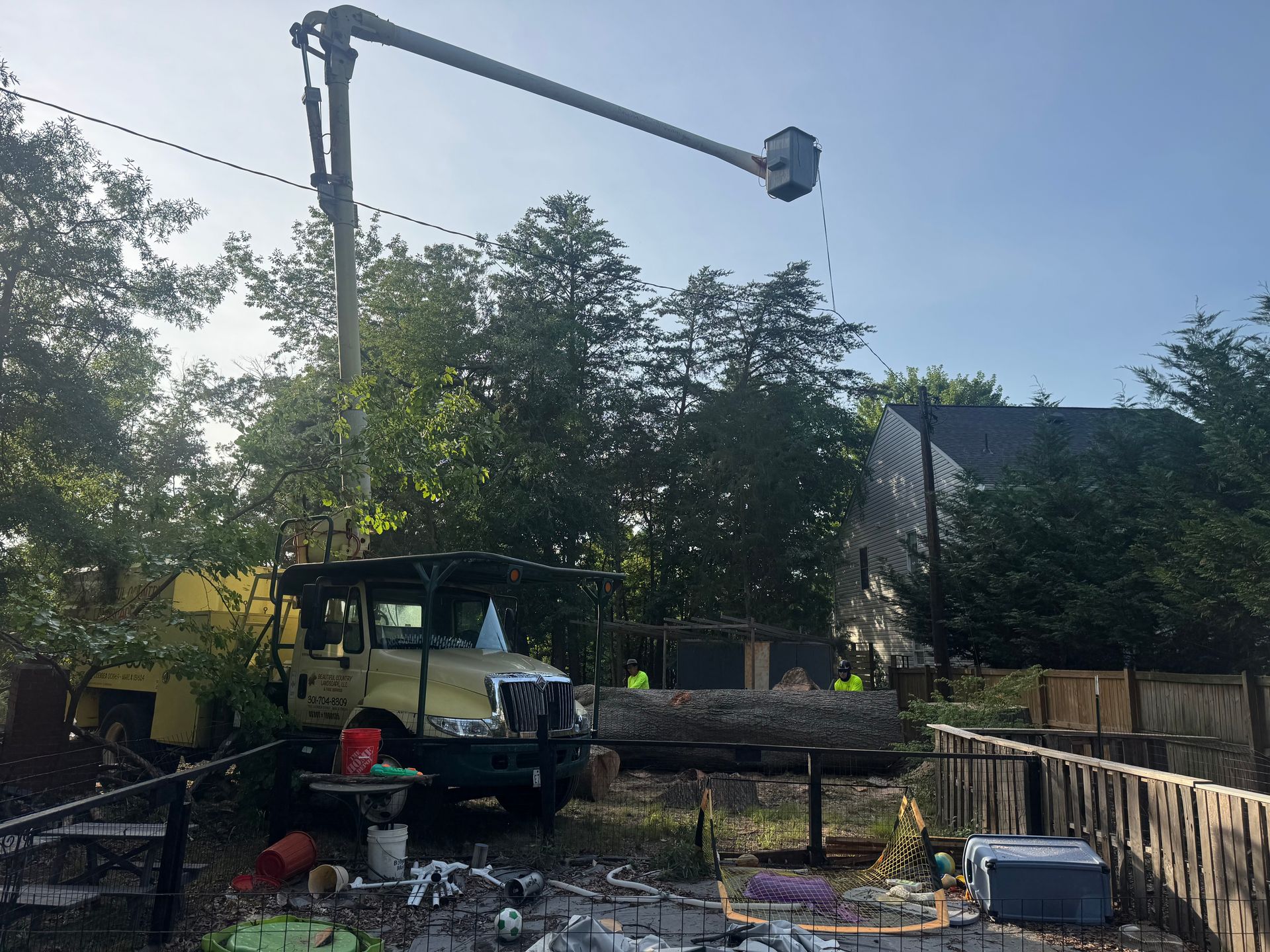 Utility truck with raised bucket arm near trees and a house in a yard, with construction materials scattered below.