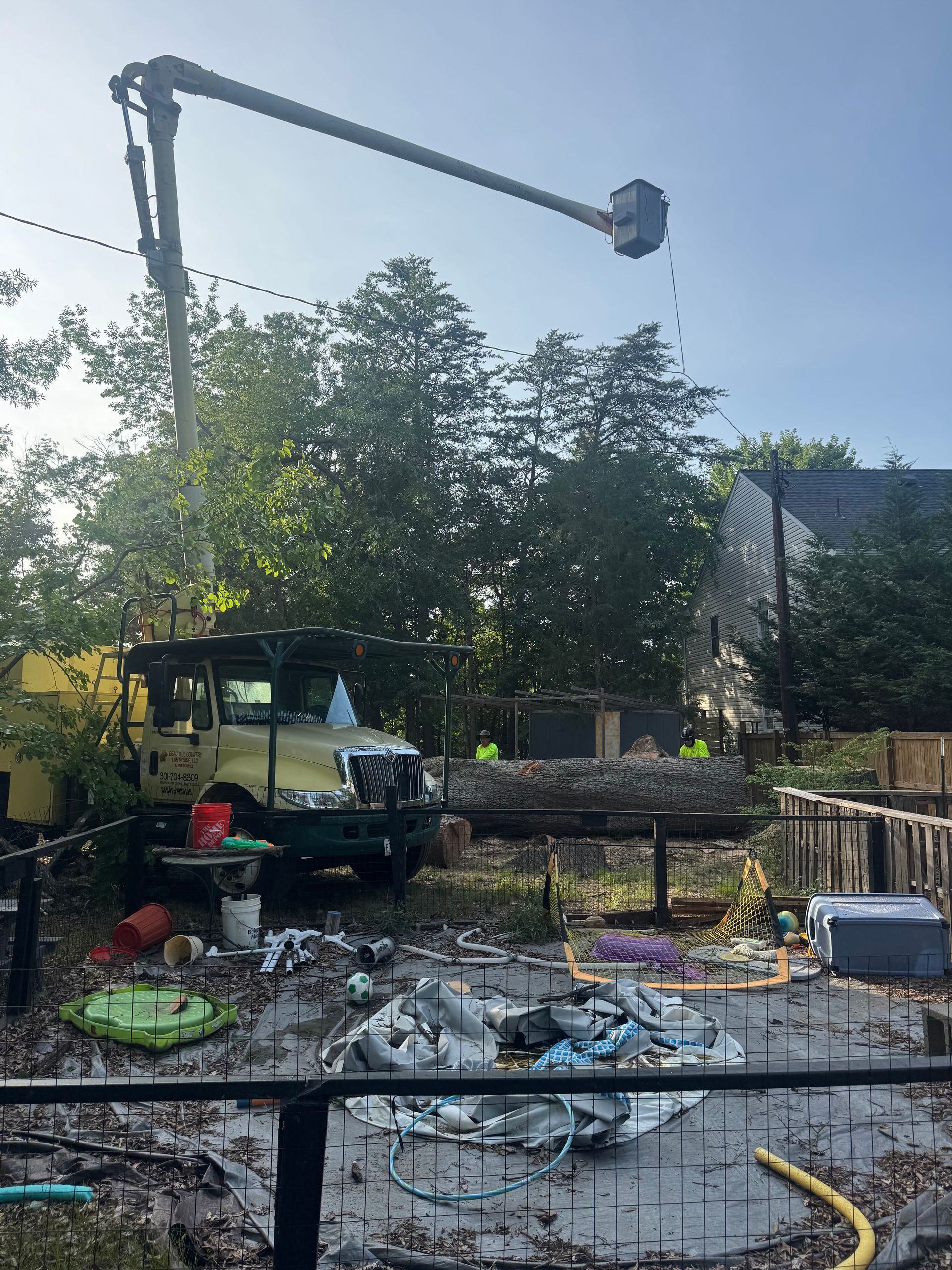Outdoor construction area with a crane arm over scattered debris and equipment under trees and blue sky