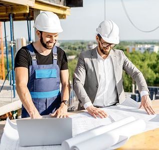 Two construction workers in hard hats review blueprints and a laptop on a building site.