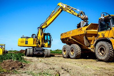 Yellow excavator loading a yellow dump truck on a construction site under a clear blue sky.