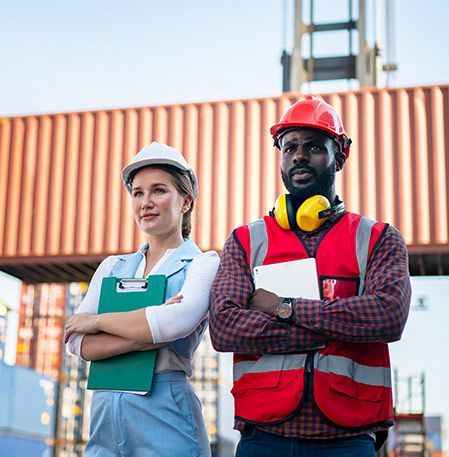 Woman and man in safety gear at a shipping port, arms crossed, looking off-camera.