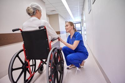 Nurse in blue scrubs kneels, talking to elderly patient in wheelchair in a hospital hallway.