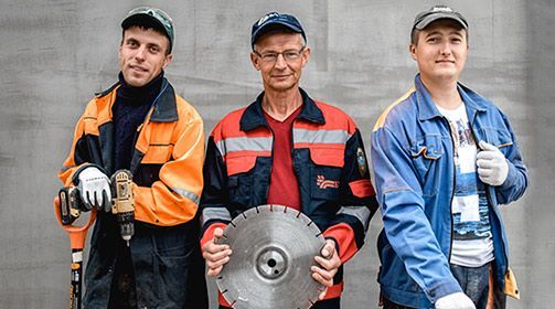 Three construction workers pose, one holding a drill, the other a saw blade, all wearing work attire.