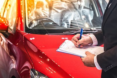 Person in suit writing on a clipboard next to a red car, likely inspecting it.