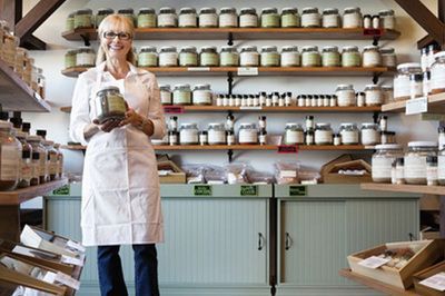 Woman in apron holds a jar in a shop with shelves of glass jars filled with products.