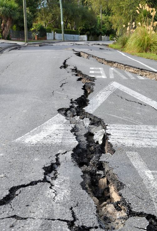 Cracked asphalt road with deep fissures, possibly from an earthquake. White crosswalk lines visible.