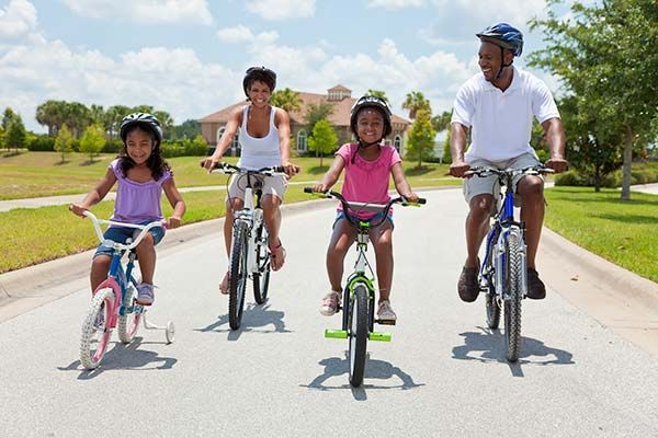 Family of four riding bikes on a paved road; sunny day, helmets worn.