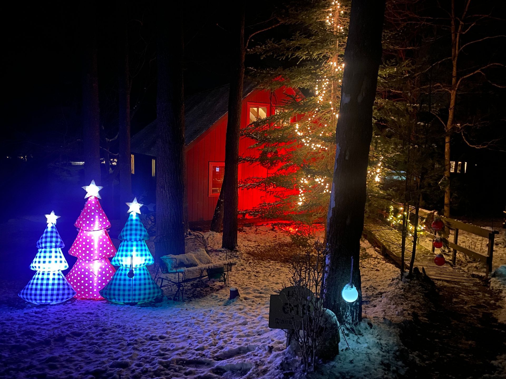 Christmas lights illuminate trees and a red house at night, with snow on the ground.
