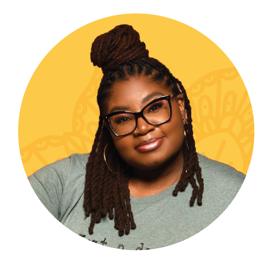 A woman with dreadlocks and glasses is smiling in front of a brick wall