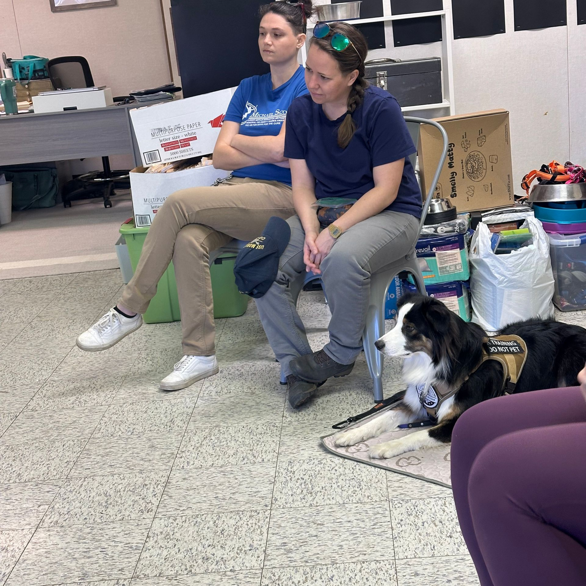 Two people sit on chairs in an office-like room, with a dog resting on the floor in front of them.