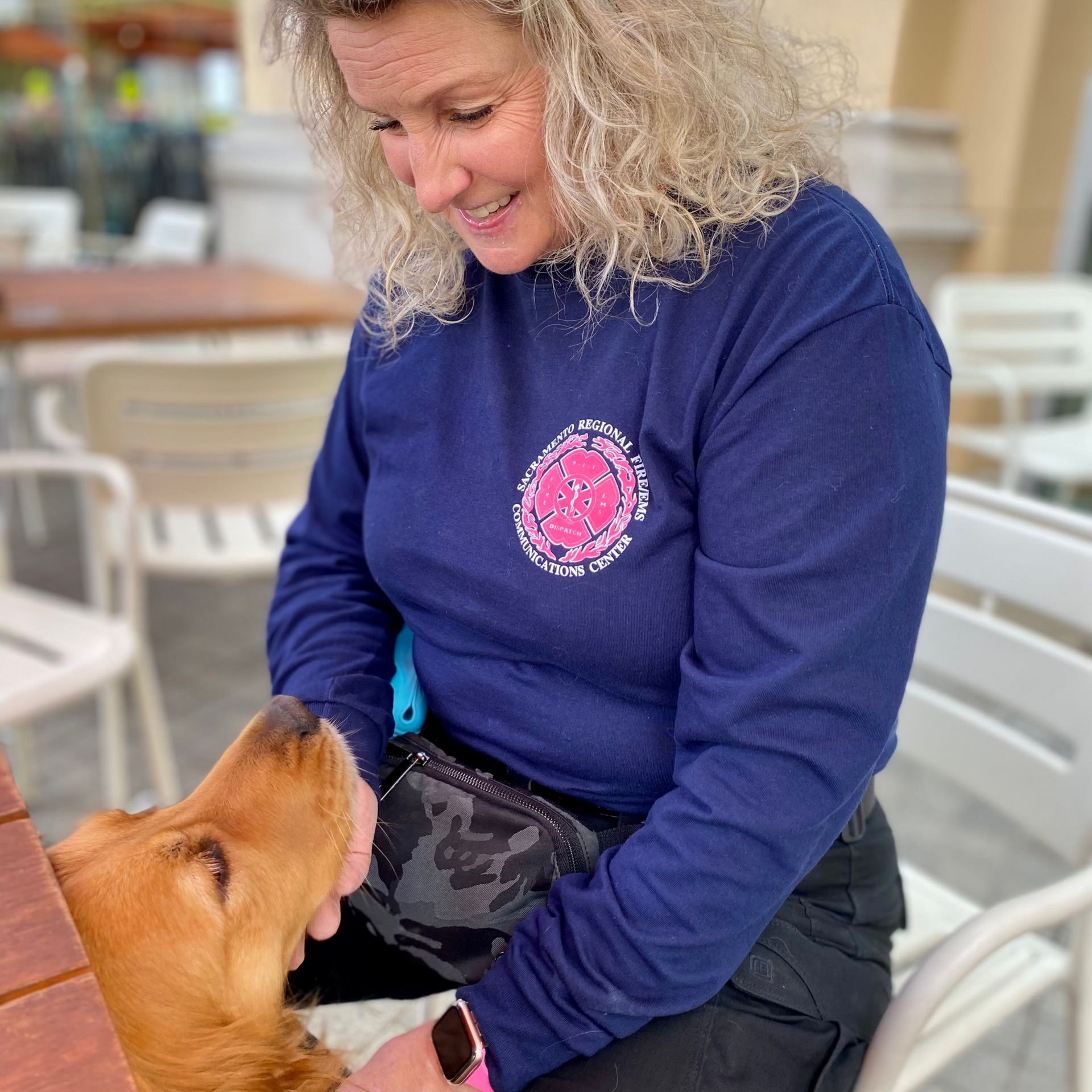 Woman in blue shirt petting a golden retriever at an outdoor table.