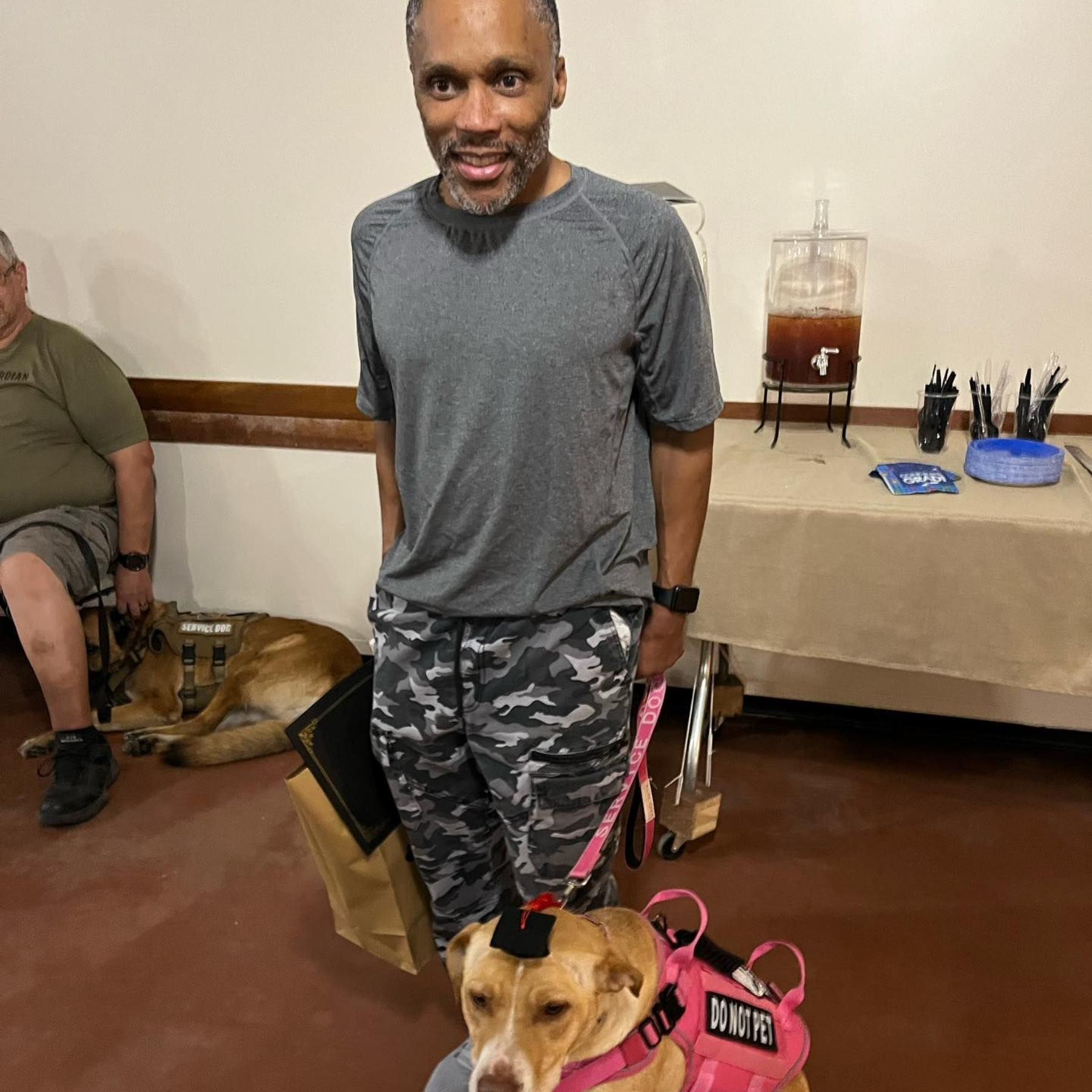 A man in a gray shirt and camouflage pants stands with a dog wearing a pink vest, inside a room with a refreshment table.