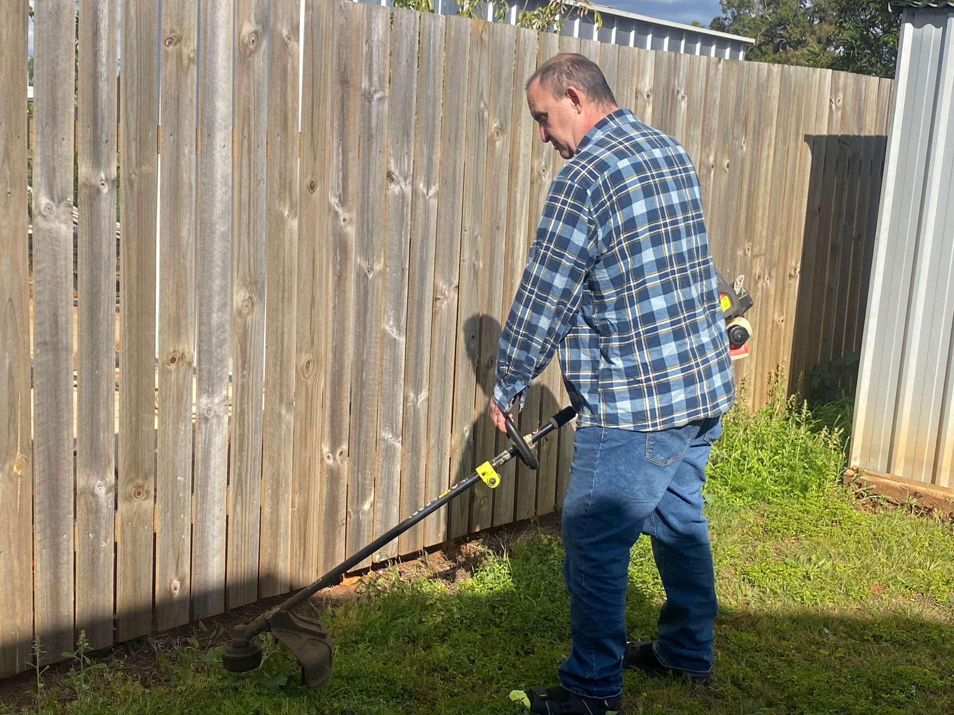 Man in a blue plaid shirt and jeans using a weed wacker on a grassy area next to a wooden fence.