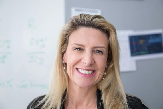 Smiling woman with blonde hair, wearing pearl earrings, against a blurry office background.