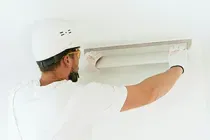 A worker in a white hard hat and gloves uses a long metal trowel to smooth plaster onto a white wall.