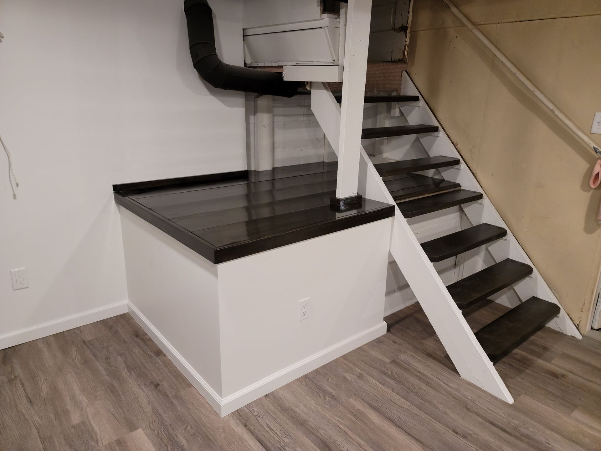 White-walled basement area with stairs, a built-in counter, and dark-colored floorboards. A black HVAC duct runs along the wall.