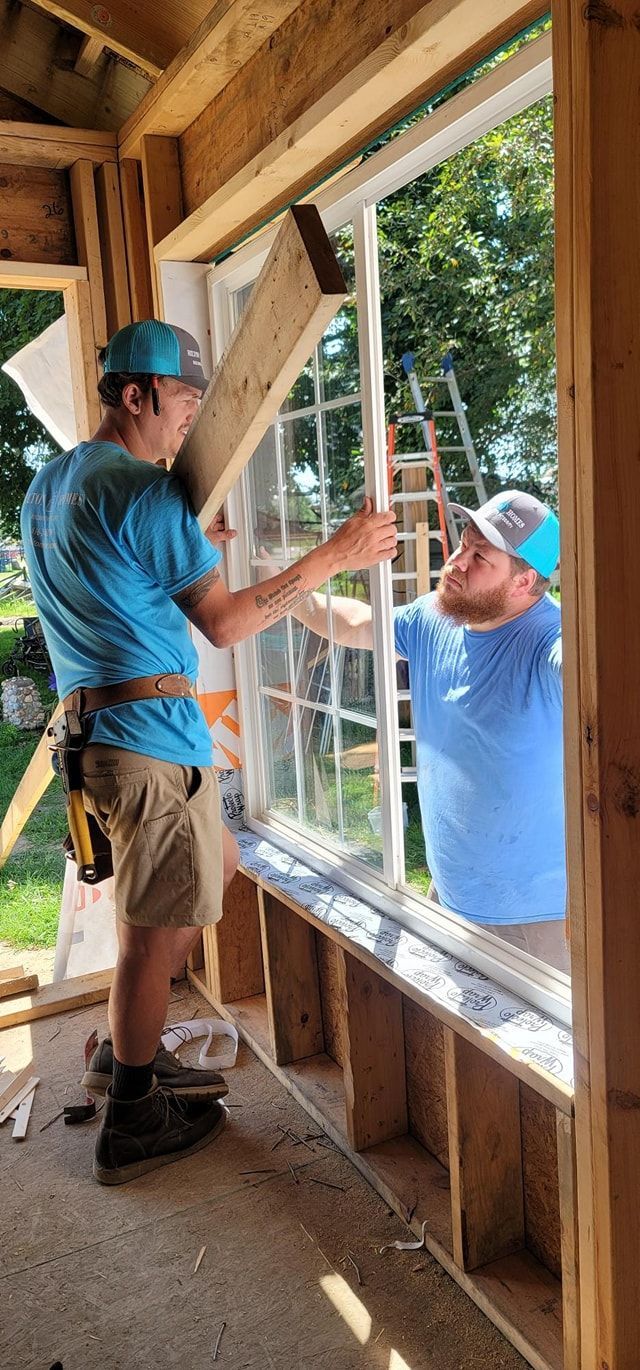 Two construction workers installing a window. One man holds a wooden piece while the other guides the window frame into place. They are wearing blue shirts and baseball caps.